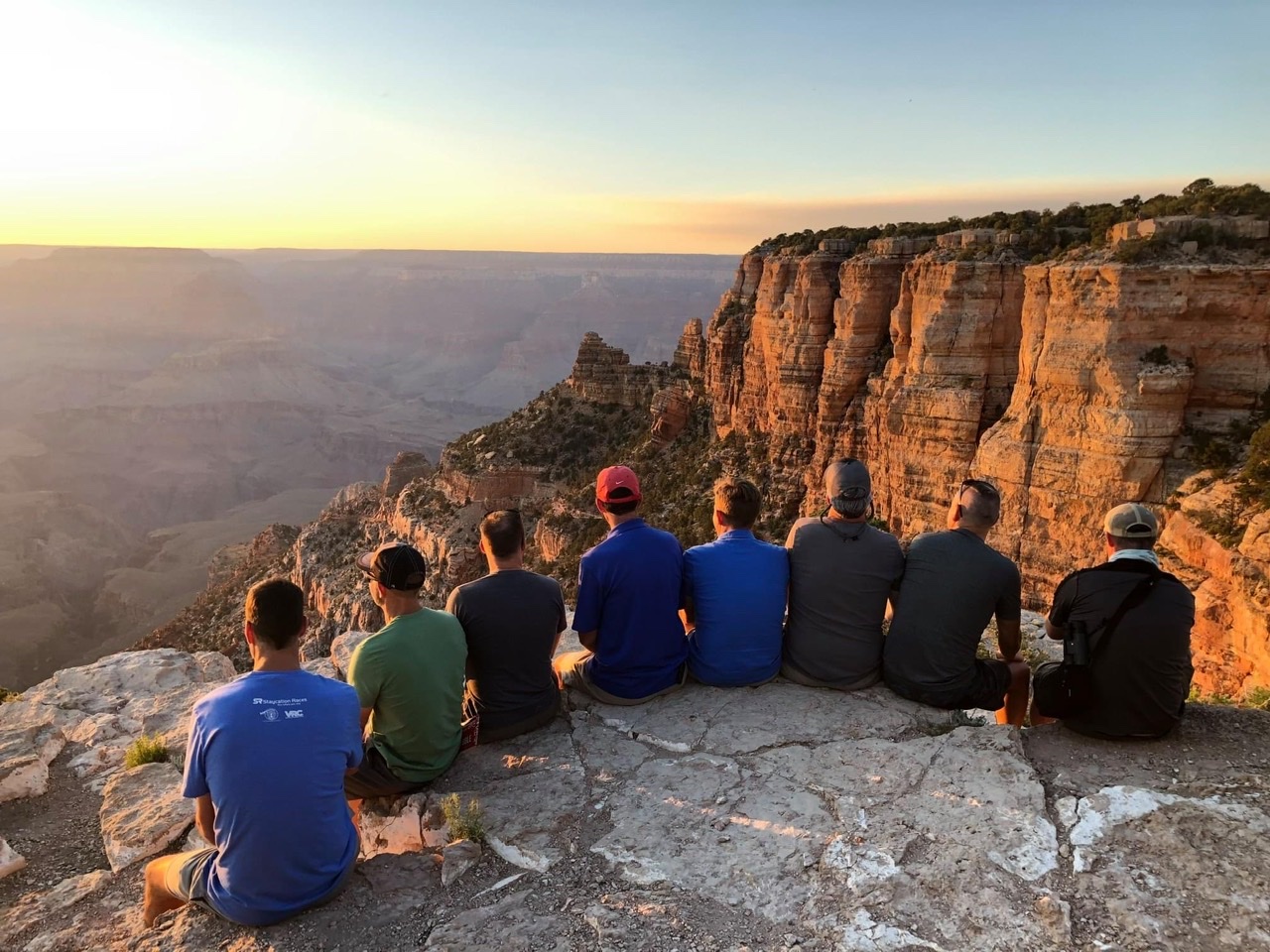 8 men sit on rock overlooking the edge of the Grand Canyon at sunset